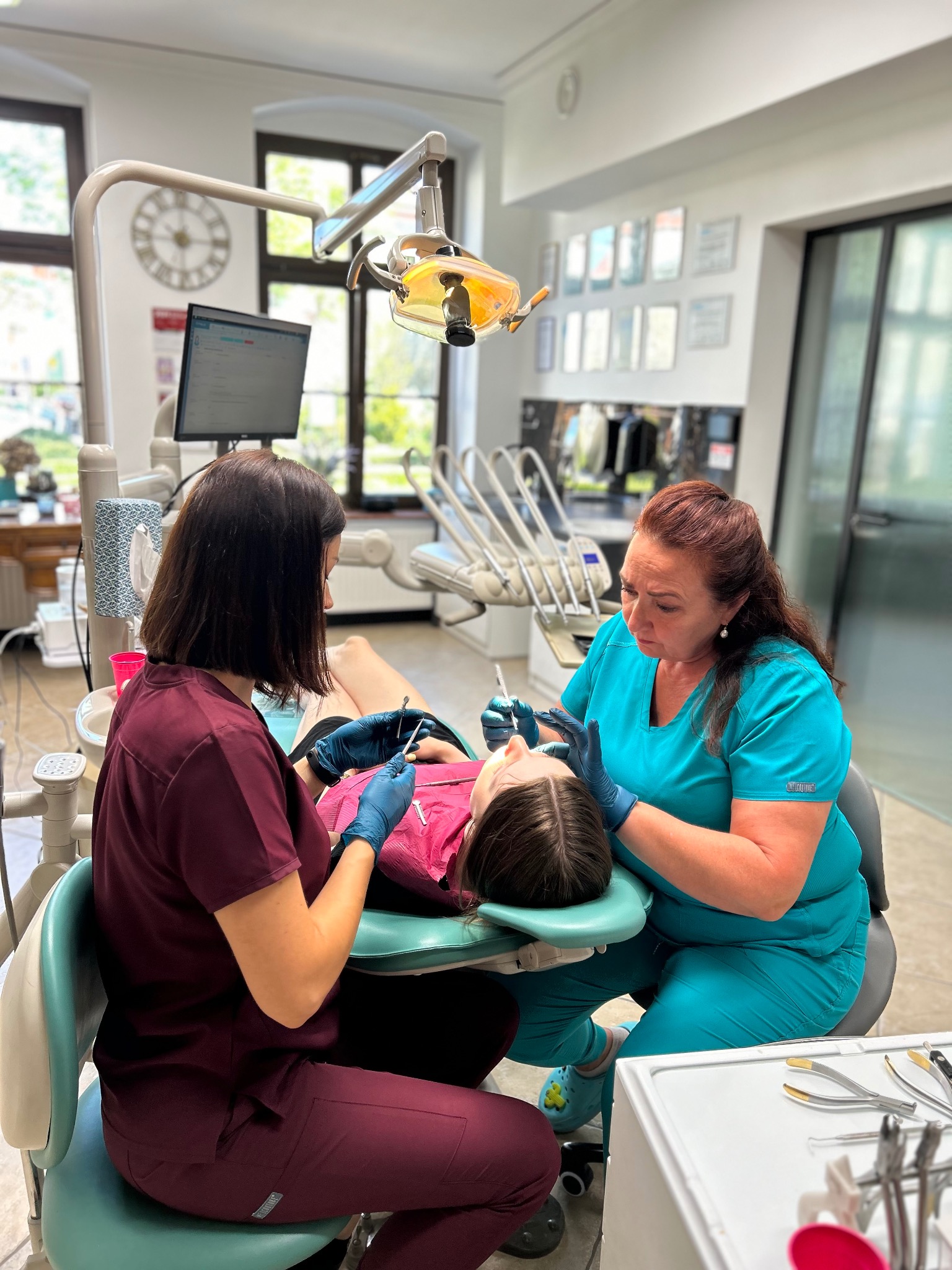 Lekarz stomatolog i asystentka w trakcie badania jamy ustnej pacjentki na fotelu dentystycznym, oświetlonym lampą zabiegową; w tle widoczny zegar i okno.