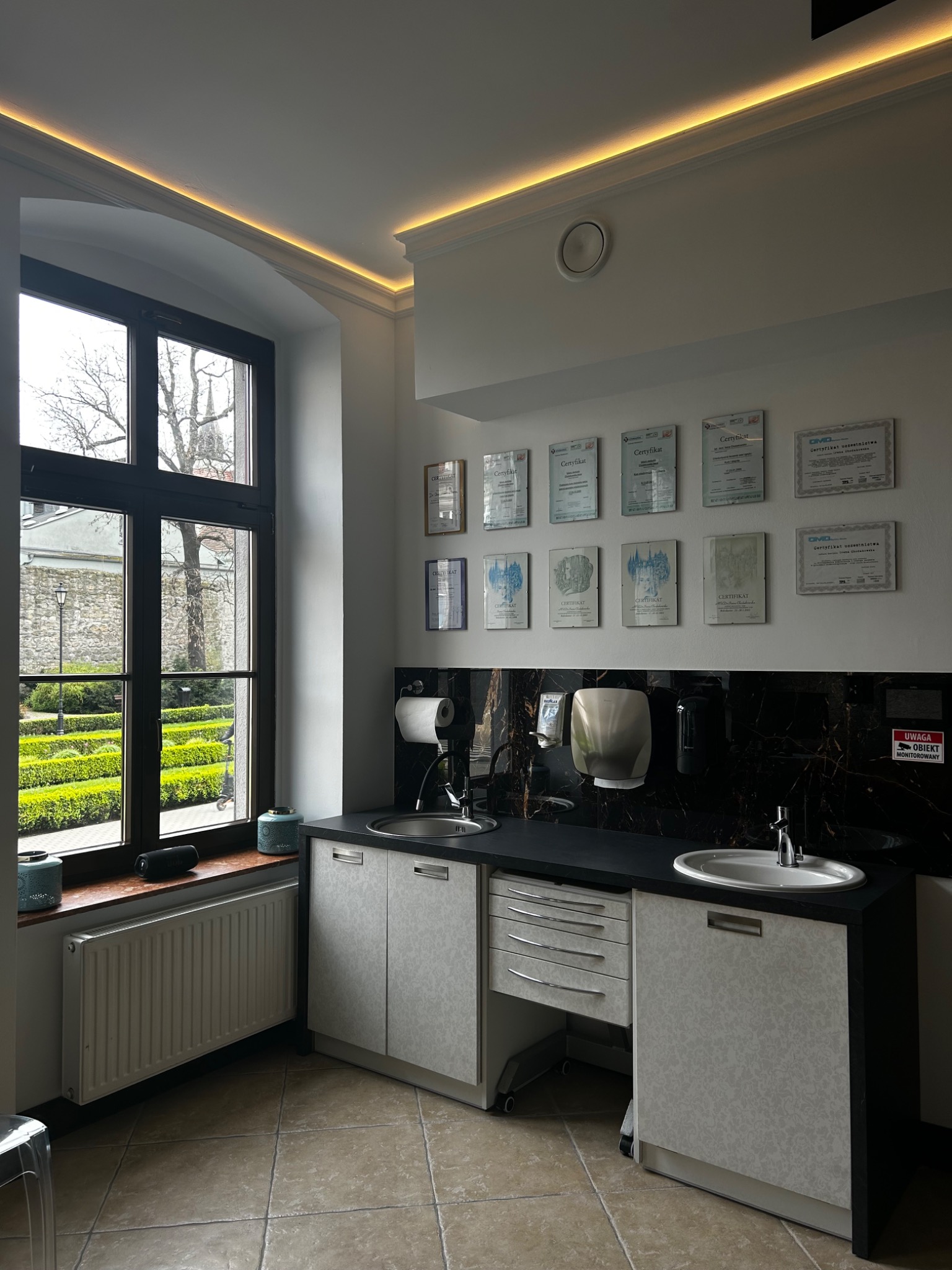 Wnętrze gabinetu stomatologicznego z oknem na park, umywalkami, szafkami i ścianą z certyfikatami, oświetleniem LED podsufitowym.
