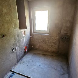 Prosta Płytka Marcin Jasiński - Surowa łazienka w trakcie przygotowania do prac wykończeniowych: ściany pokryte szarym tynkiem, widoczne instalacje wodno-kanalizacyjne, sufit z wiszącą żarówką, okno z białymi ramami i poziomica...
