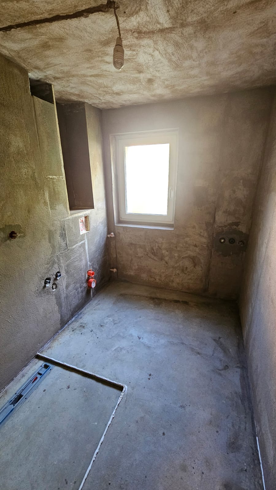 Surowa łazienka w trakcie przygotowania do prac wykończeniowych: ściany pokryte szarym tynkiem, widoczne instalacje wodno-kanalizacyjne, sufit z wiszącą żarówką, okno z białymi ramami i poziomica...