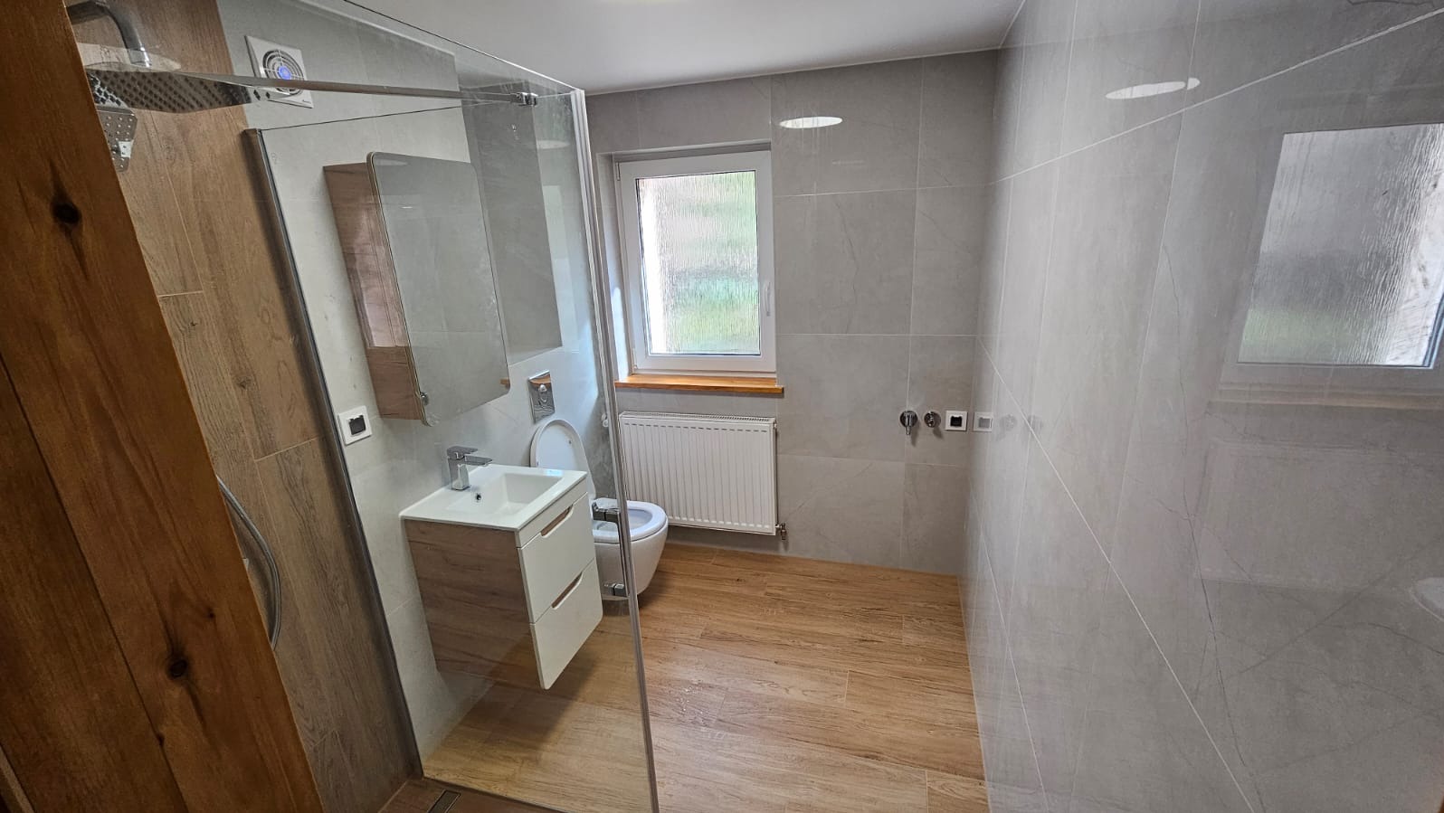 Nowoczesna łazienka z drewnopodobną podłogą, szarą glazurą ścienną, kabiną prysznicową typu walk-in i wiszącą szafką z umywalką.