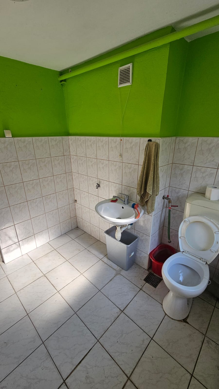 Łazienka z zielonymi ścianami i białą glazurą przed remontem, widoczna umywalka z szarą szafką, toaleta z otwartą klapą i ręcznik na ścianie.