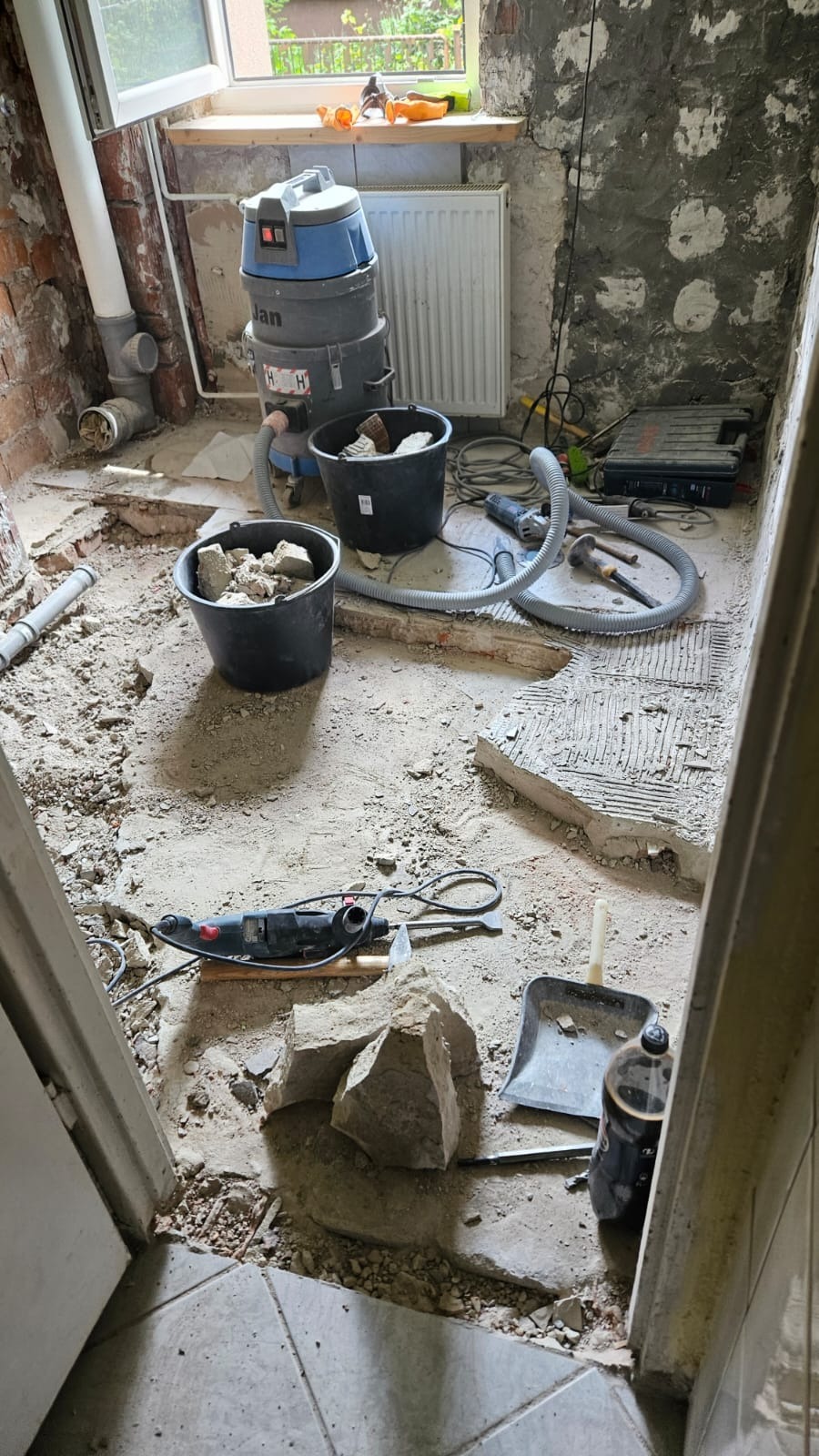 Wnętrze łazienki we Wrocławiu w trakcie rozbiórki, widoczne dwa wiadra z gruzem, odkurzacz przemysłowy, narzędzia i fragmenty starej glazury.