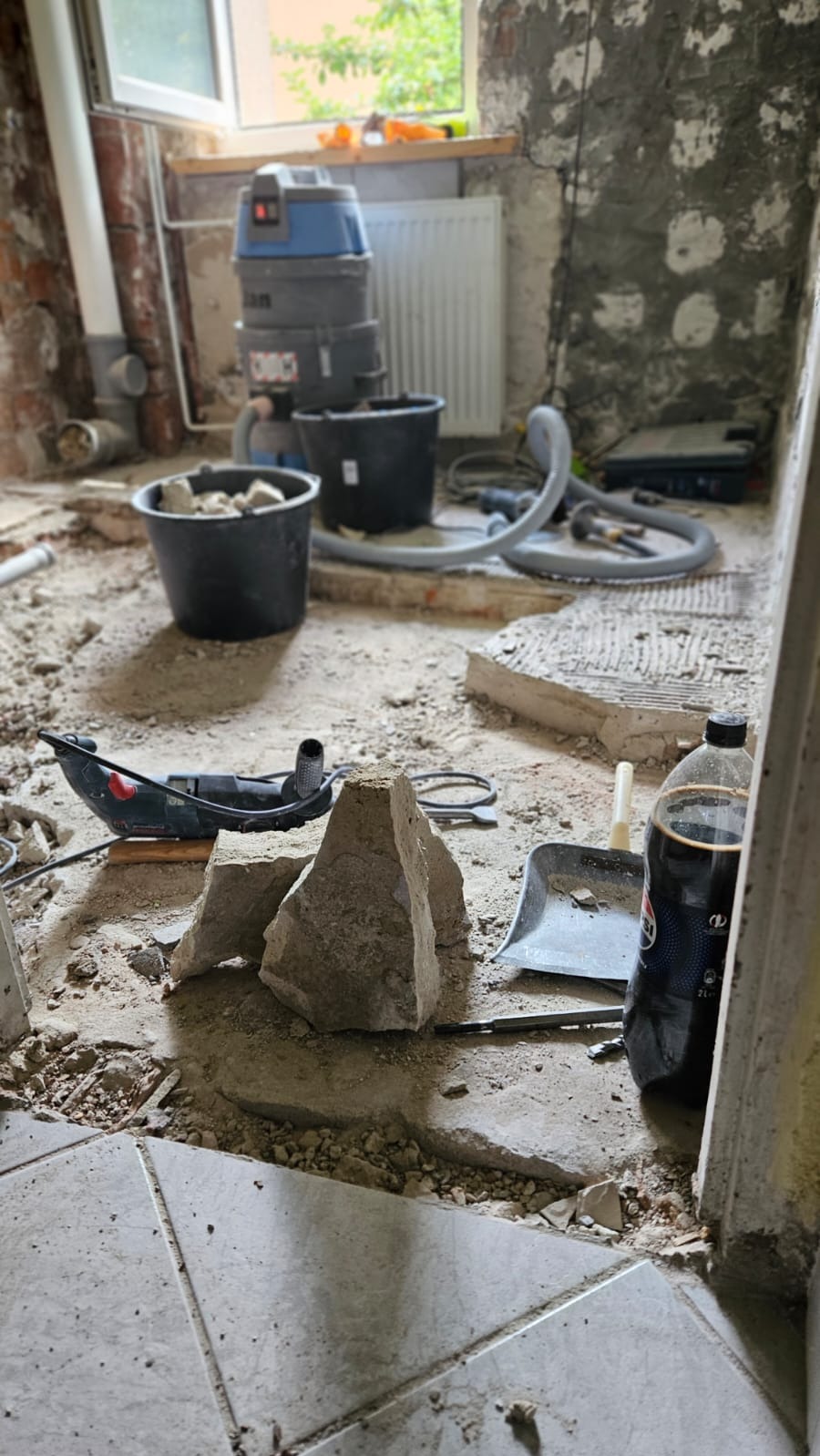 Wnętrze łazienki w trakcie rozbiórki: widoczne gruz, odkurzacz przemysłowy, narzędzia, wiadro z resztkami i butelka napoju przy framudze drzwi.