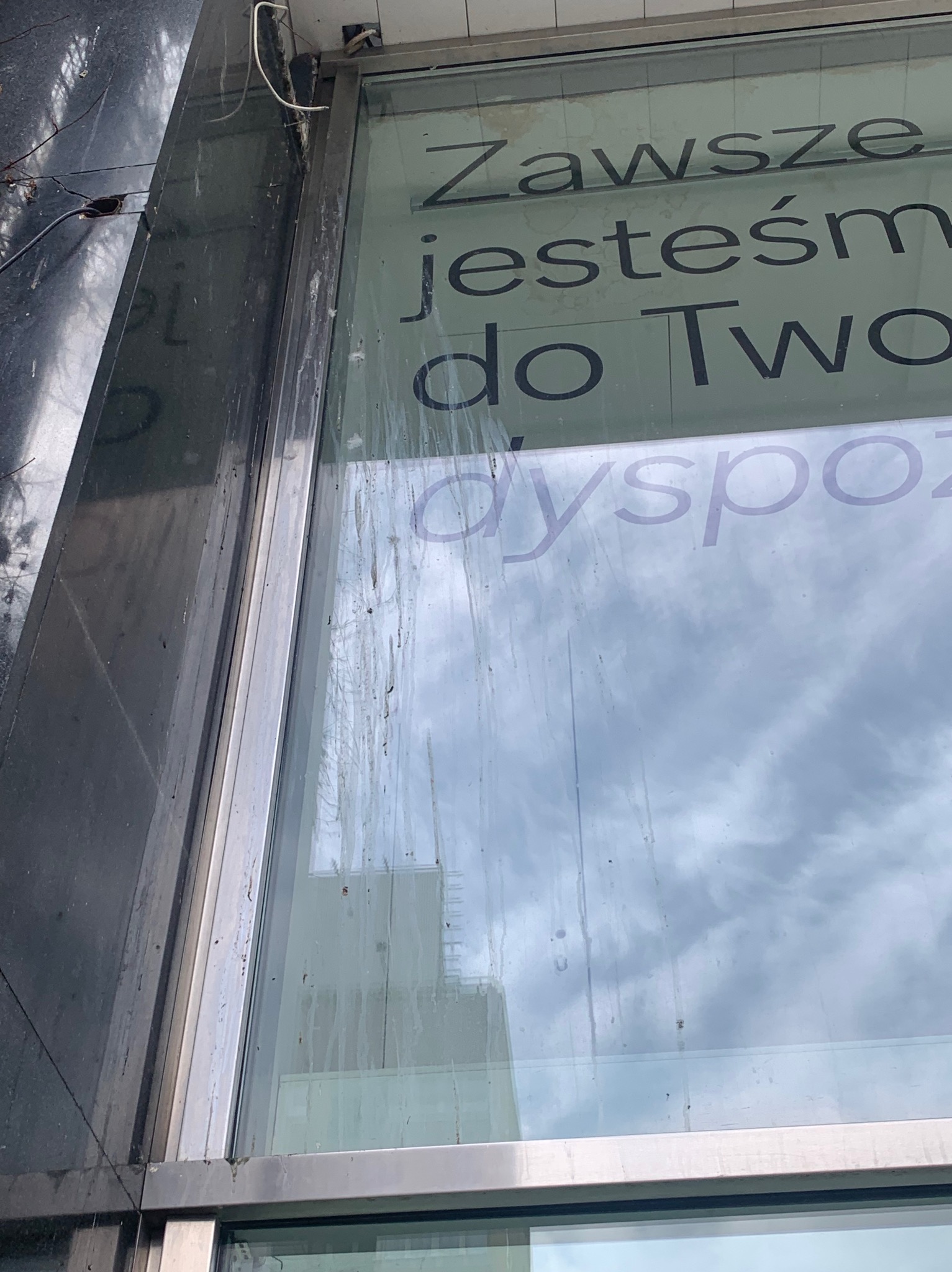 Brudna szyba okienna z napisem 'Zawsze jestem do Twojej dyspozycji', widoczne zacieki i zabrudzenia na szkle, odblask chmur.