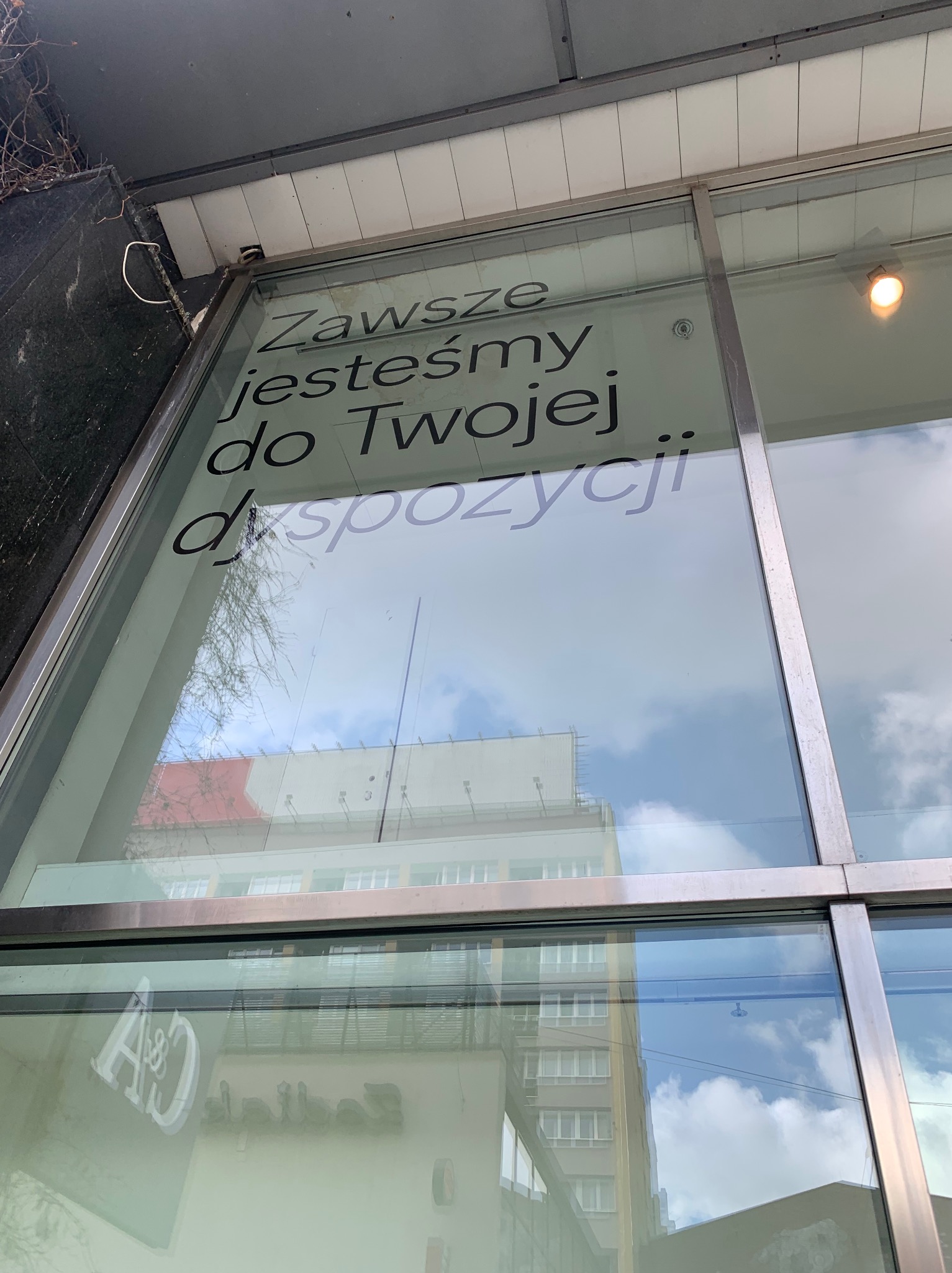 Okno z napisem 'Zawsze jesteśmy do Twojej dyspozycji' i odbiciem budynku oraz nieba, widoczne elementy elewacji i instalacji.