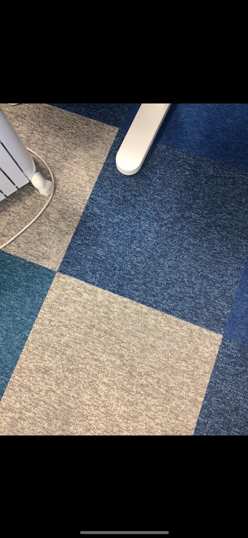 Fragment biura z kwadratową wykładziną dywanową w odcieniach szarości i błękitu oraz białą nogą biurka i grzejnikiem.