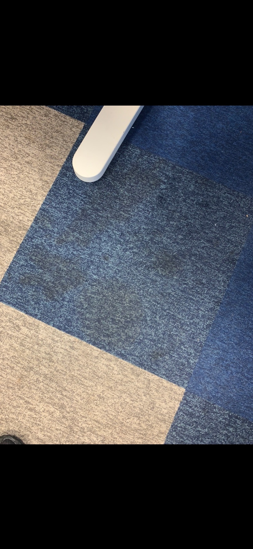 Fragment biurowej podłogi pokrytej kwadratowymi płytami dywanowymi w odcieniach szarości i błękitu, widoczne ślady miejscowego zabrudzenia na niebieskim dywanie, obok fragment białej nogi od biurka.