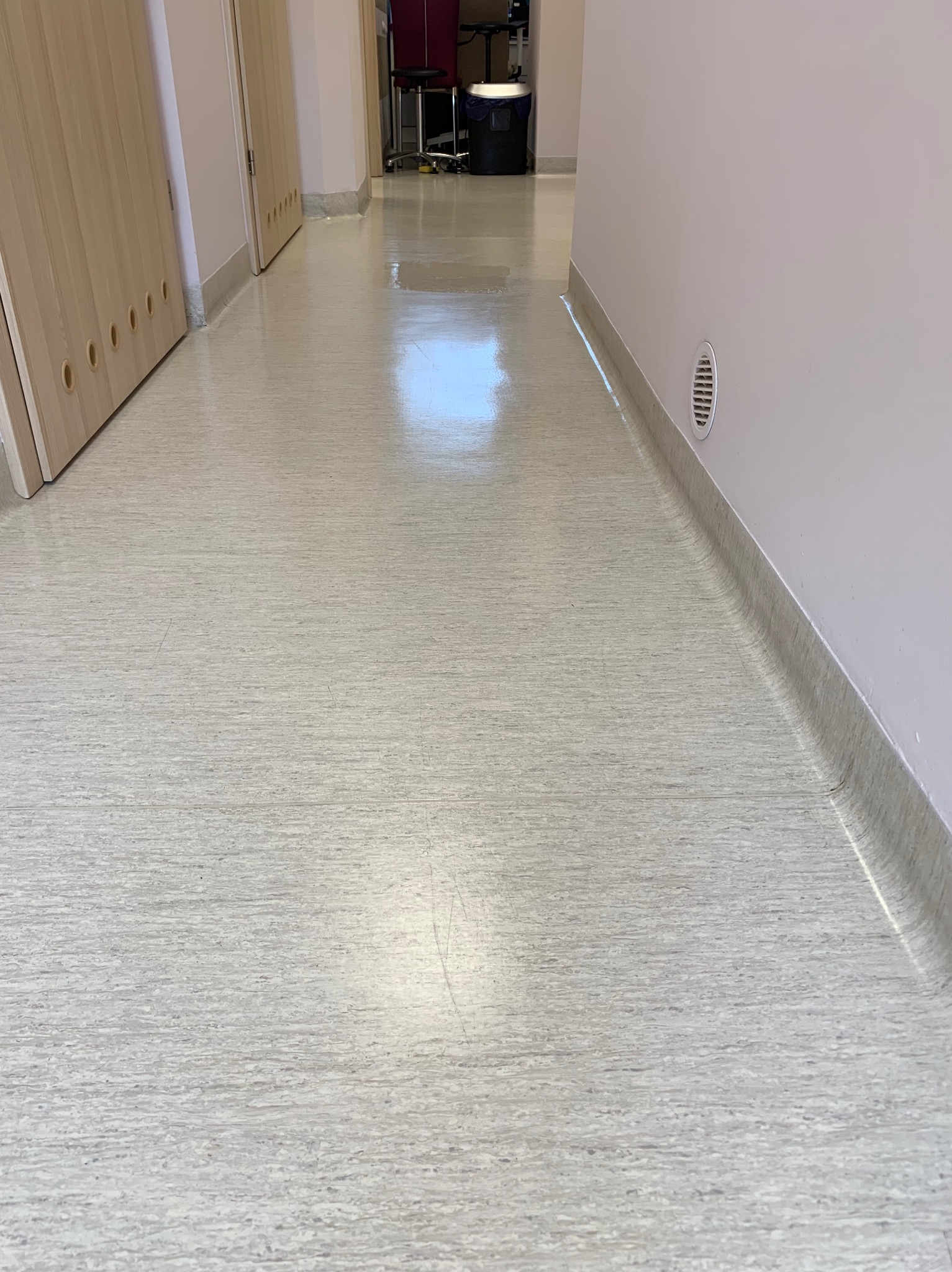 Błyszcząca, jasna podłoga korytarza w budynku biurowym, widoczne drzwi po lewej stronie i wentylacja w ścianie po prawej.