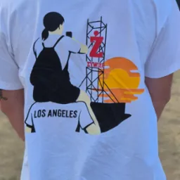 Biała koszulka z unikalnym nadrukiem: sylwetka kobiety, wieża z logo, napis 'Los Angeles' i motyw zachodzącego słońca. Widok z tyłu, na zewnątrz.