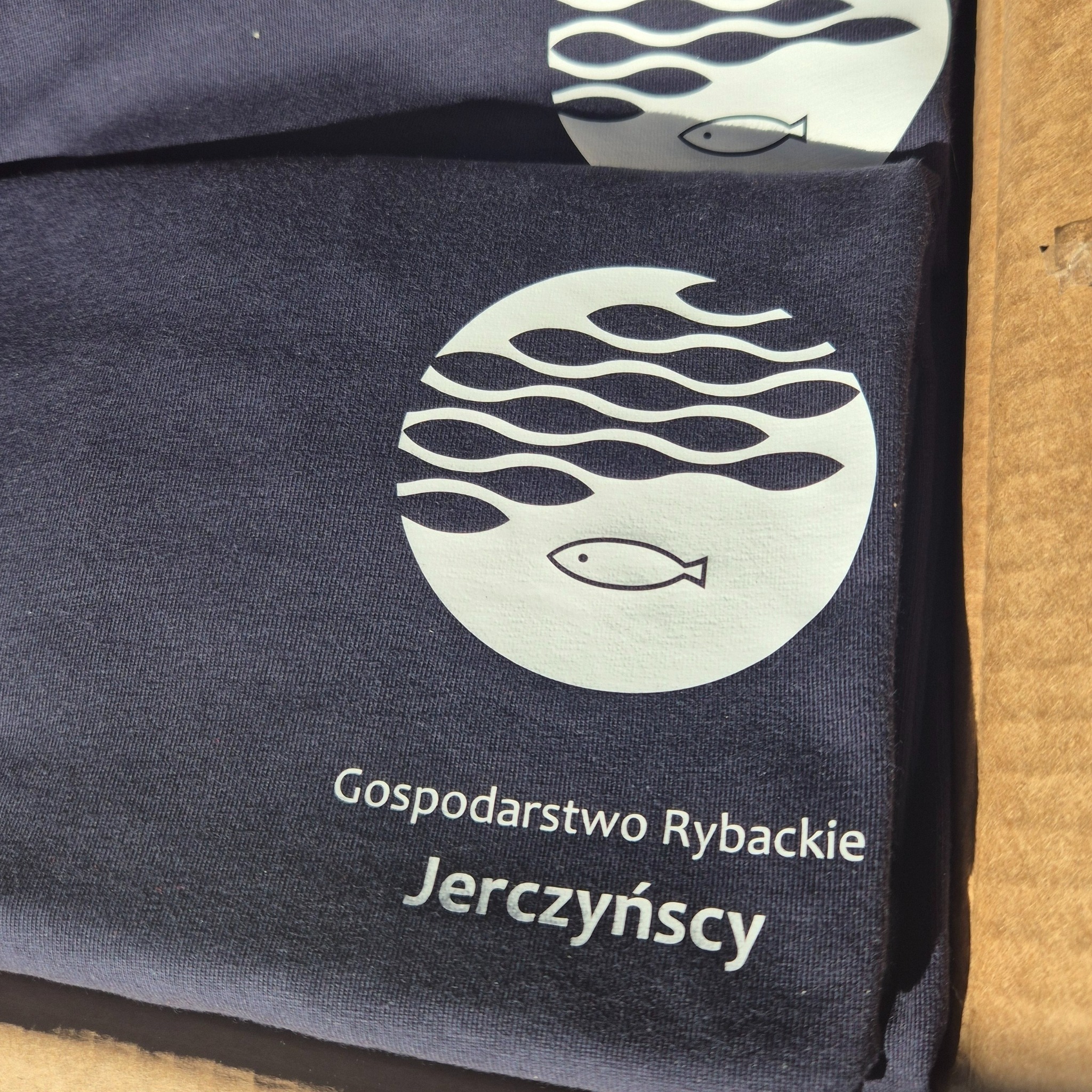 Stos granatowych koszulek z białym nadrukiem logo firmy 'Gospodarstwo Rybackie Jerczyńscy', widocznych kilka sztuk w kartonowym pudełku.