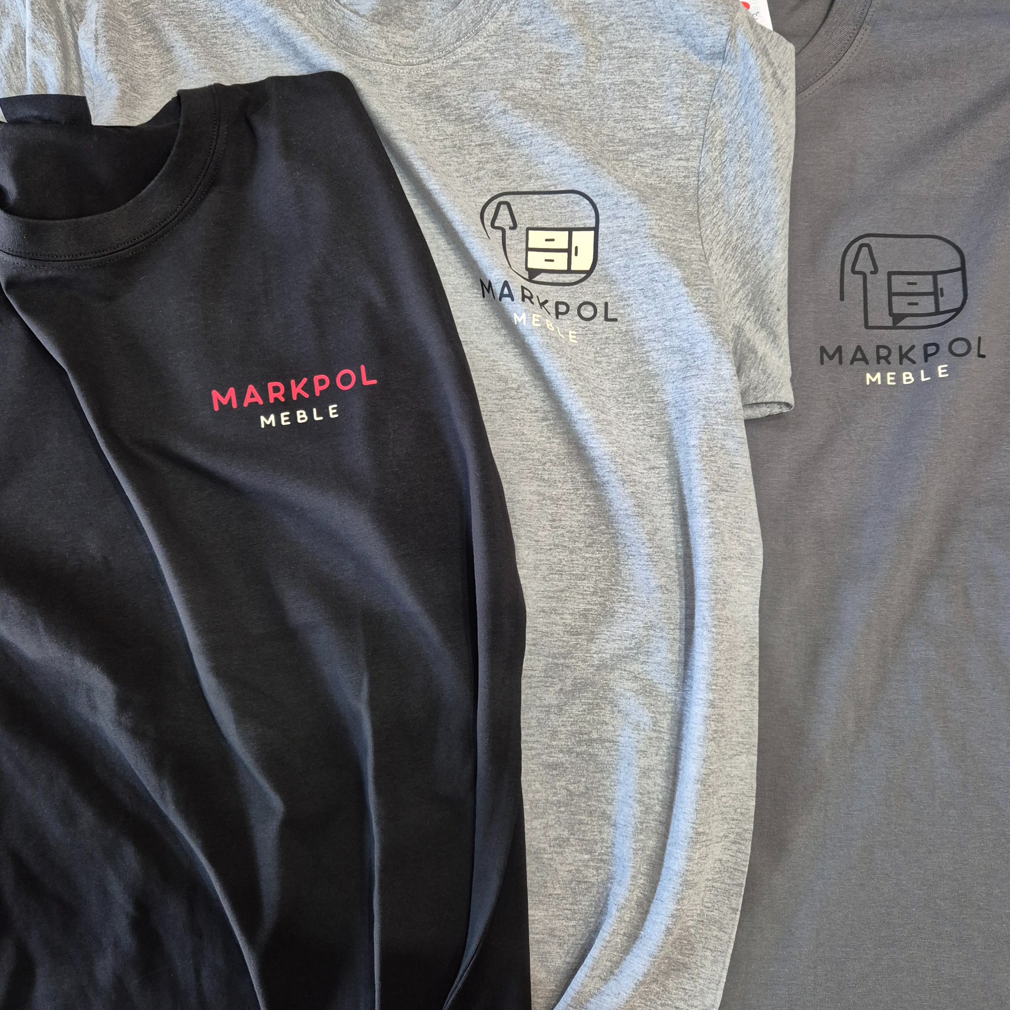 Trzy koszulki z krótkim rękawem w kolorach: czarnym, szarym i grafitowym, z nadrukowanym logo firmy meblowej 'MARKPOL MEBLE', prezentowane z bliska.