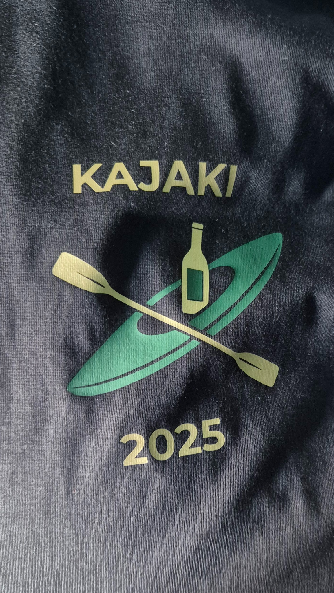 Niebieska koszulka z nadrukiem 'Kajaki 2025', przedstawiającym kajak, wiosło i butelkę. Nadruk w odcieniach zieleni i żółci. Widoczna faktura materiału.