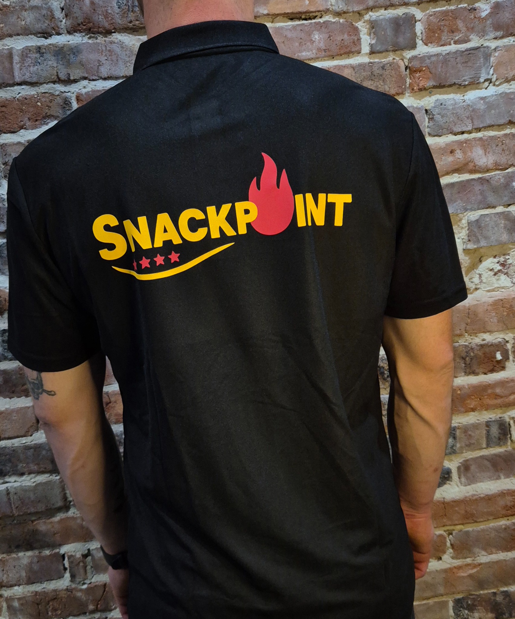 Czarna koszulka polo z żółto-czerwonym nadrukiem 'SNACKPOINT' i trzema gwiazdkami, widok z tyłu na tle ceglanej ściany, prezentacja nadruku na odzieży.