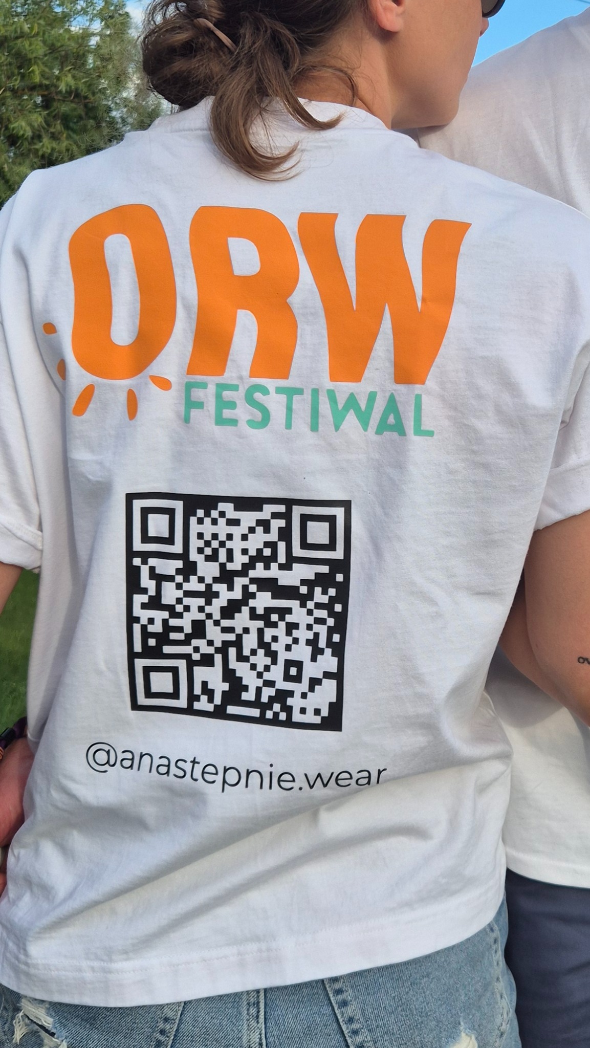 Biała koszulka z pomarańczowym napisem 'ORW', zielonym 'Festiwal', kodem QR i adresem @anastepnie.wear, noszona przez osobę z widocznym fragmentem jeansów.