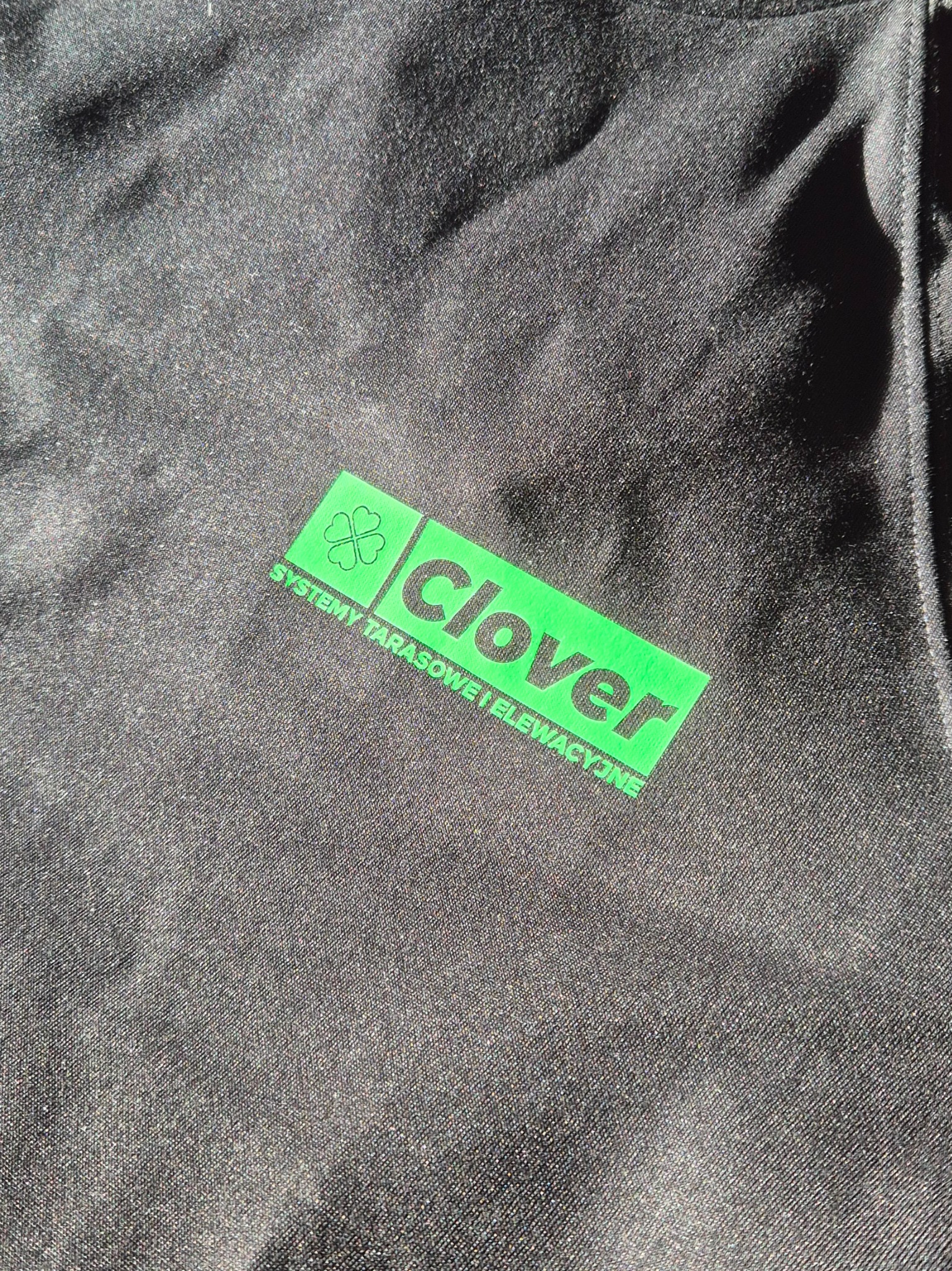 Czarna koszulka z zielonym nadrukiem logo Clover i napisem 'Systemy tarasowe i elewacyjne', widoczna faktura materiału.
