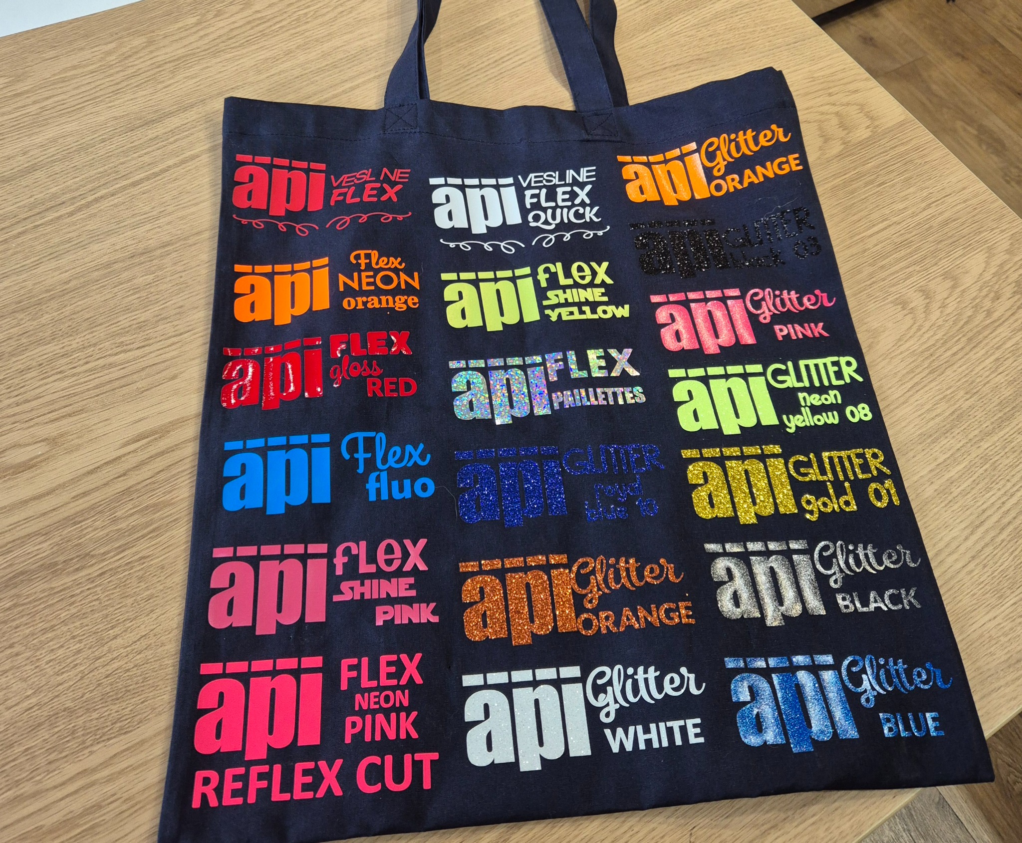 Czarna torba bawełniana z kolorowymi nadrukami API Flex, prezentująca różne techniki i efekty, takie jak glitter, neon, shine, na drewnianym stole.