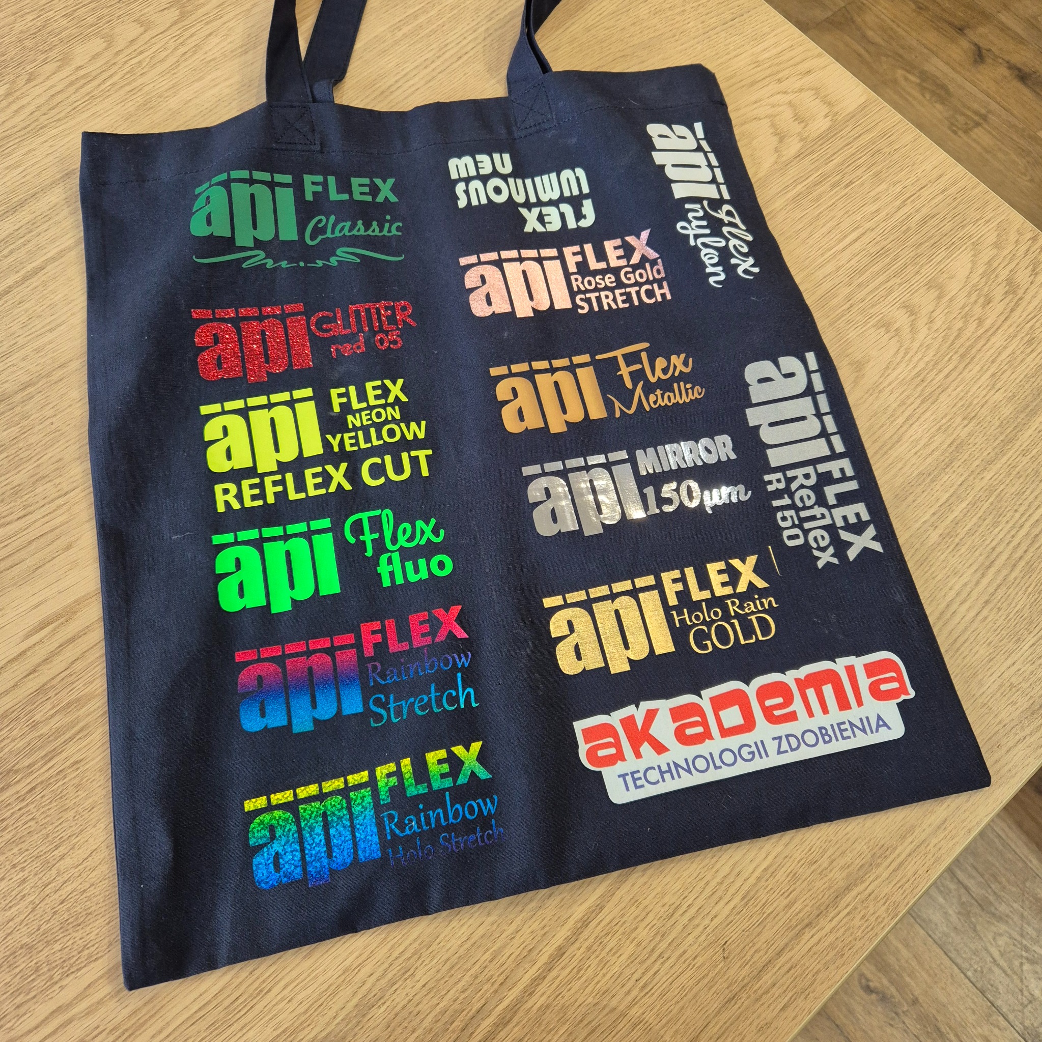 Czarna torba z nadrukami folii flex w różnych kolorach i wzorach, prezentująca techniki zdobienia odzieży, leżąca na drewnianym stole.