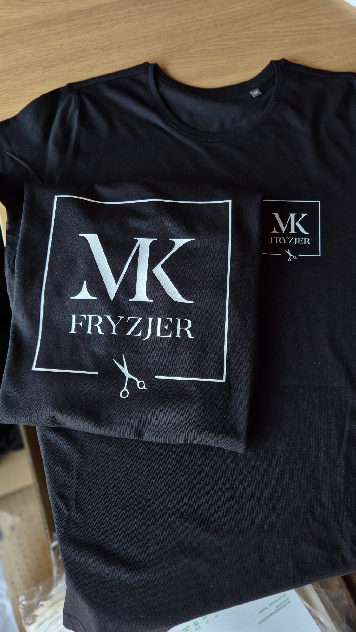 Czarne koszulki z białym nadrukiem 'MK Fryzjer' i symbolem nożyczek, leżące na drewnianym stole. Widoczne logo w dwóch rozmiarach.