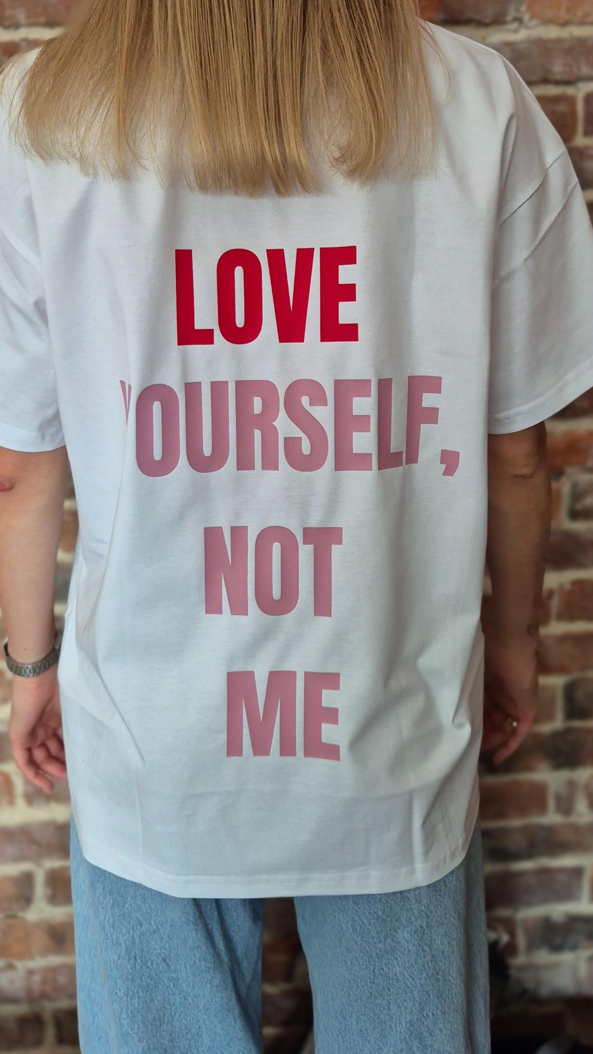 Biała koszulka z nadrukiem 'LOVE YOURSELF, NOT ME' w odcieniach różu i czerwieni, noszona przez osobę stojącą tyłem na tle ceglanej ściany. Widoczne jasne włosy i fragment dżinsów.