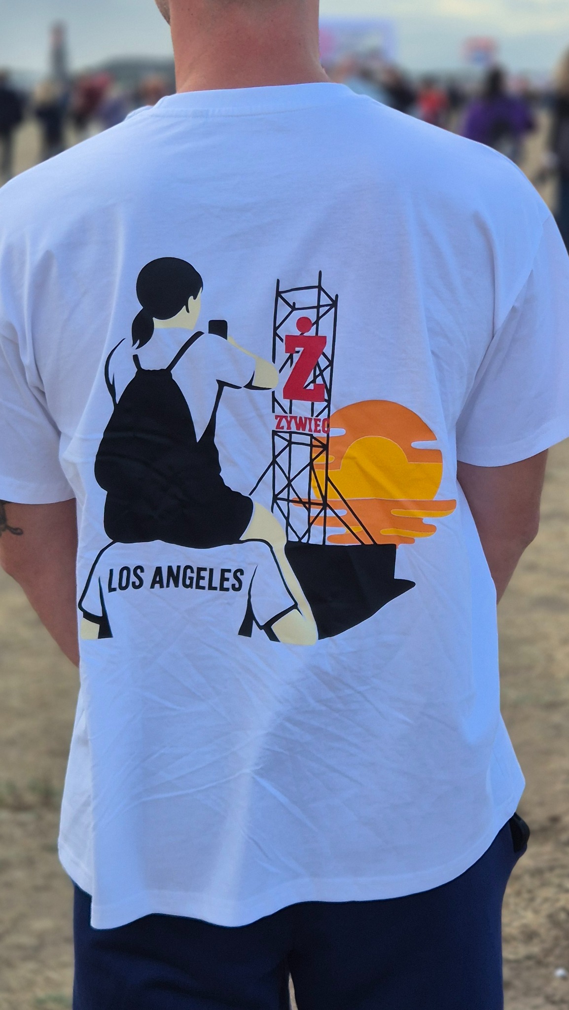 Biała koszulka z unikalnym nadrukiem: sylwetka kobiety, wieża z logo, napis 'Los Angeles' i motyw zachodzącego słońca. Widok z tyłu, na zewnątrz.