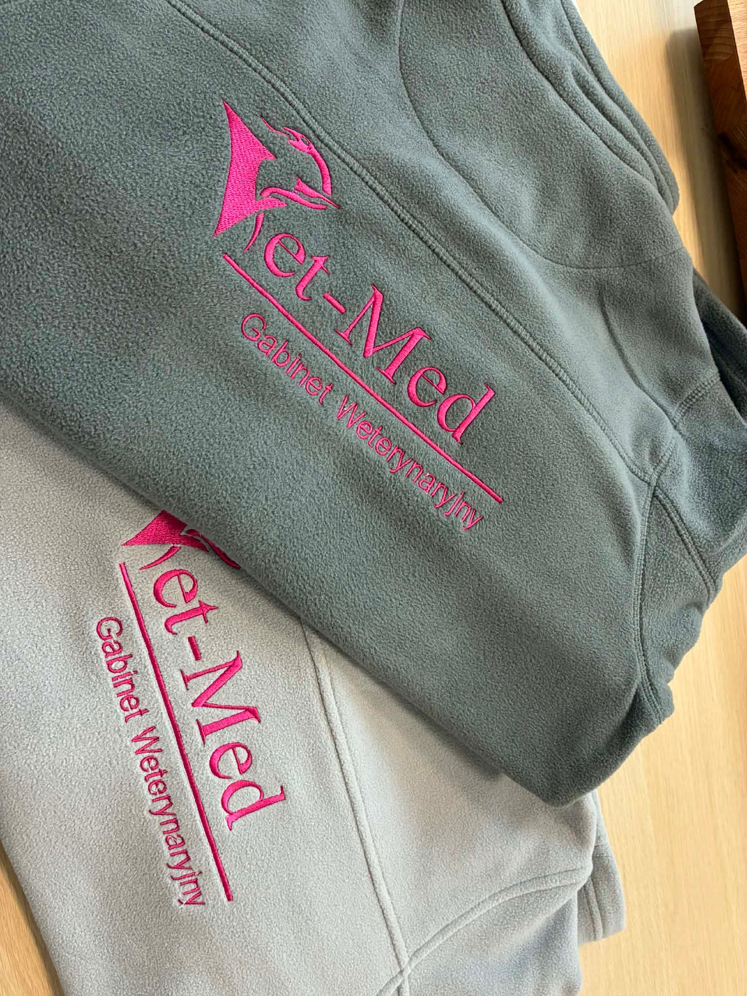 Dwie polarowe bluzy w odcieniach szarości z różowym haftem 'Vet-Med Gabinet Weterynaryjny', leżące na drewnianym blacie. Widoczne detale materiału i precyzja wykonania haftu.