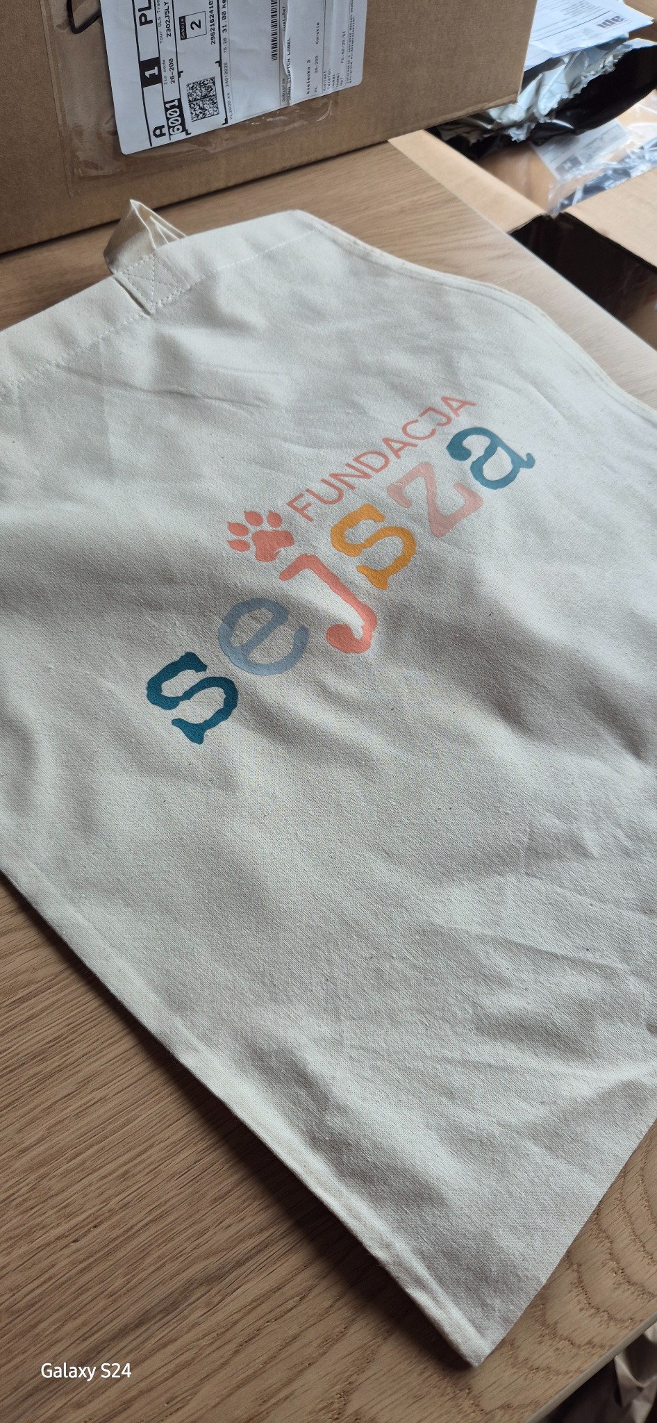 Beżowa torba bawełniana z kolorowym nadrukiem 'Fundacja Sejsza' i motywem łapy, leżąca na drewnianym stole. Widoczne pudełka w tle.