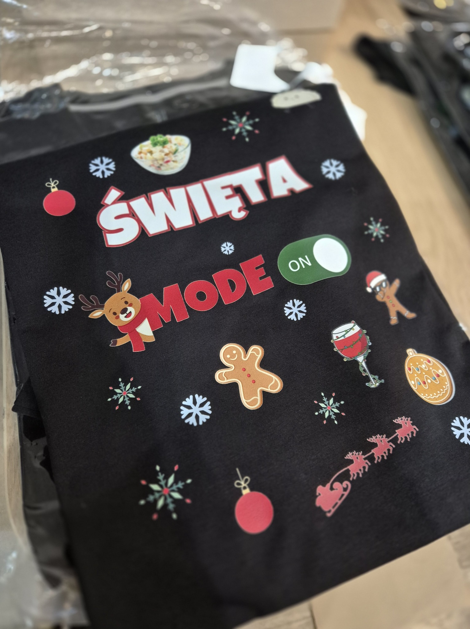 Czarna koszulka z nadrukiem 'Święta MODE ON', motywami świątecznymi: sałatka, renifer, pierniki, bombki, sanie Mikołaja. Widoczne opakowanie foliowe.