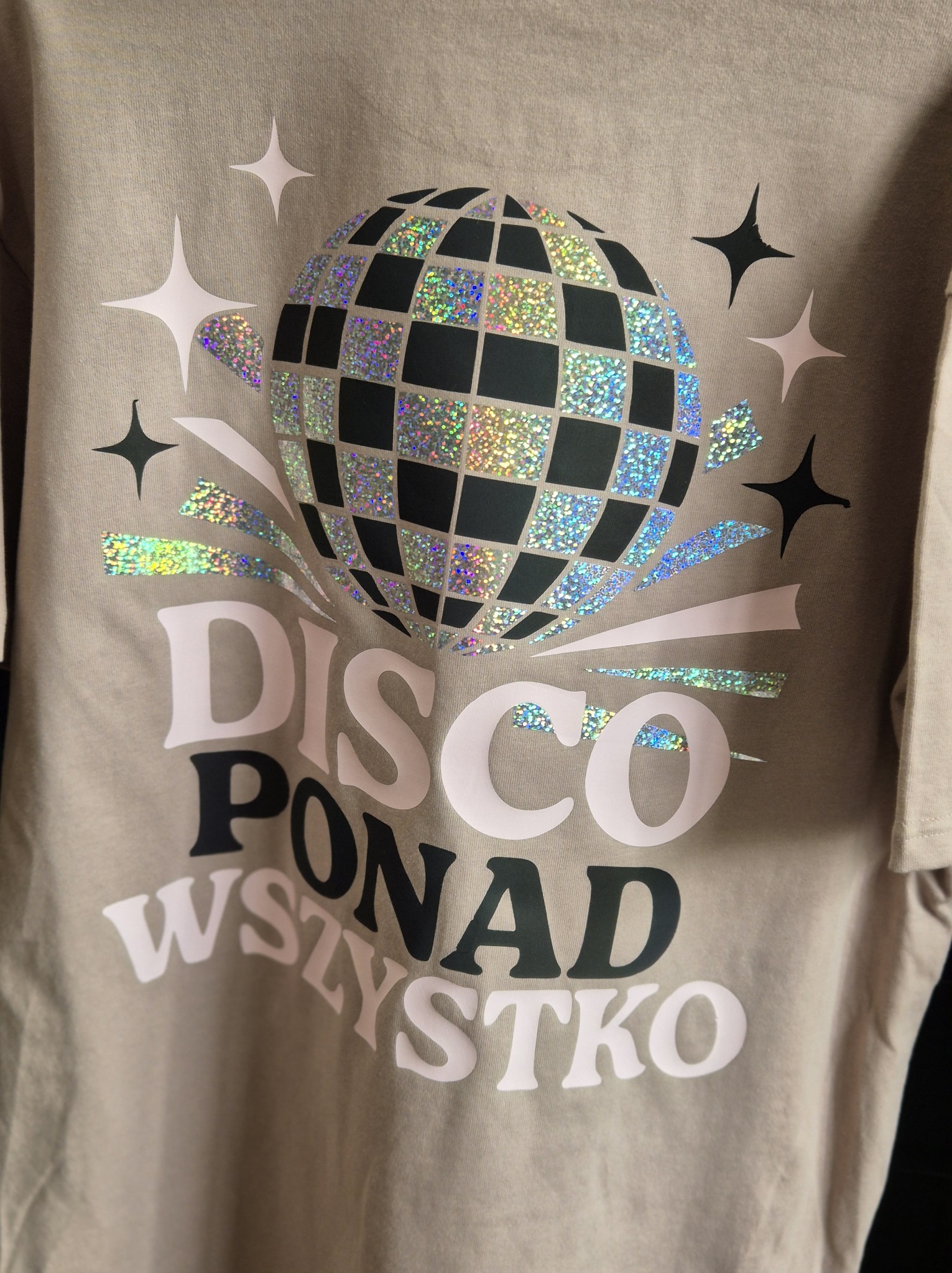 Beżowa koszulka z błyszczącym nadrukiem w stylu disco: kula, gwiazdy i napis 'Disco ponad wszystko'.