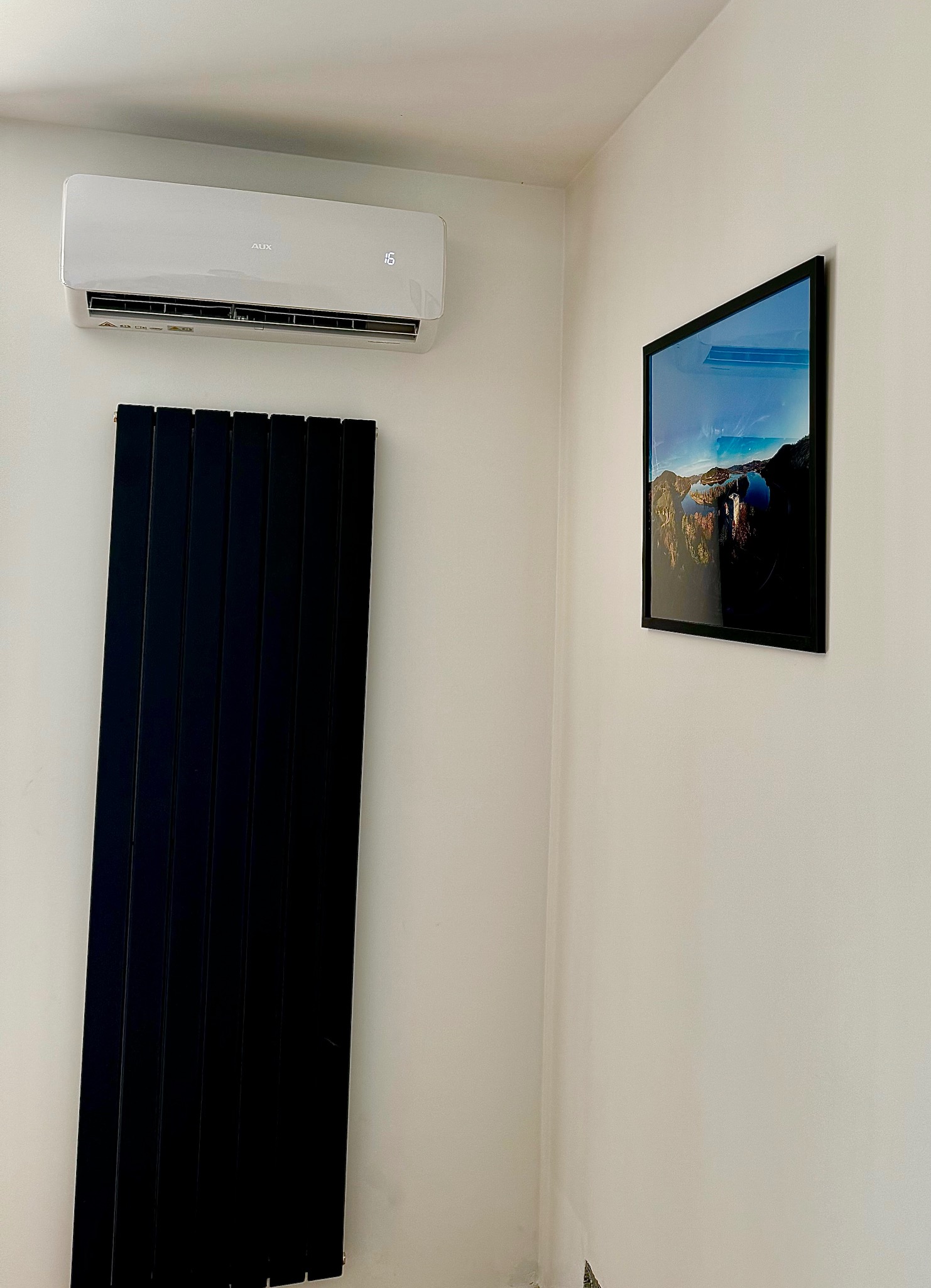 Biała klimatyzacja AUX zamontowana na ścianie w pomieszczeniu, obok czarny grzejnik panelowy i obraz w czarnej ramie.