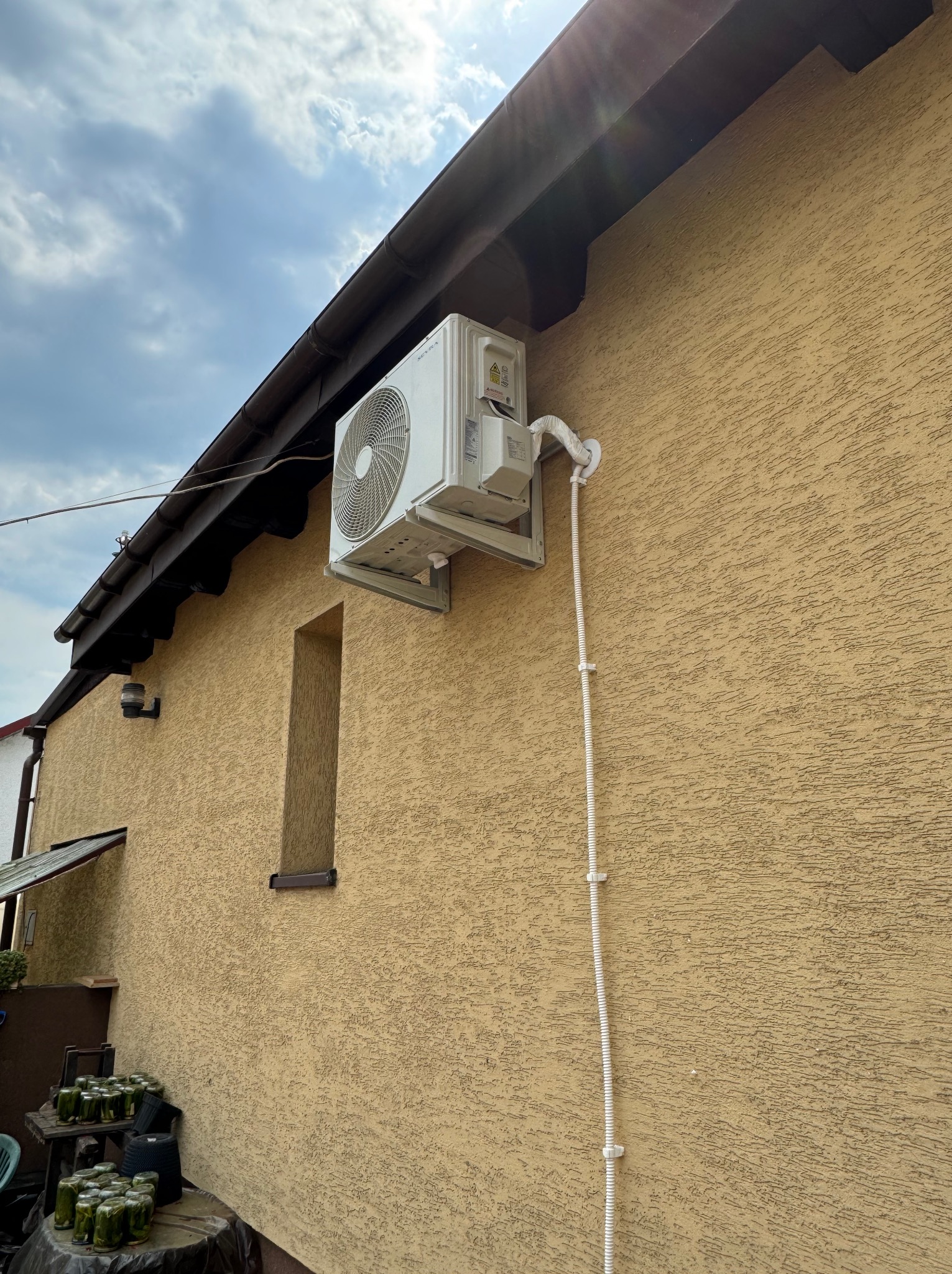 Zewnętrzna jednostka klimatyzacji zamontowana na elewacji budynku z widocznymi przewodami i uchwytami montażowymi. W tle widoczne słoiki z ogórkami kiszonymi.