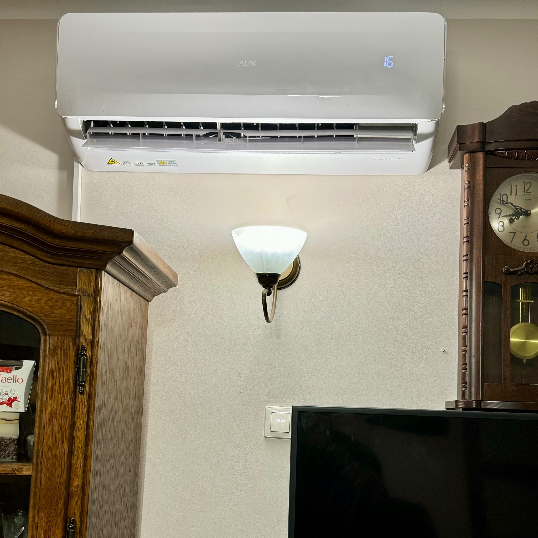 Biały klimatyzator AUX zamontowany na ścianie w pomieszczeniu z elementami klasycznego wystroju, w tle widoczny zegar kominkowy i drewniana szafka.
