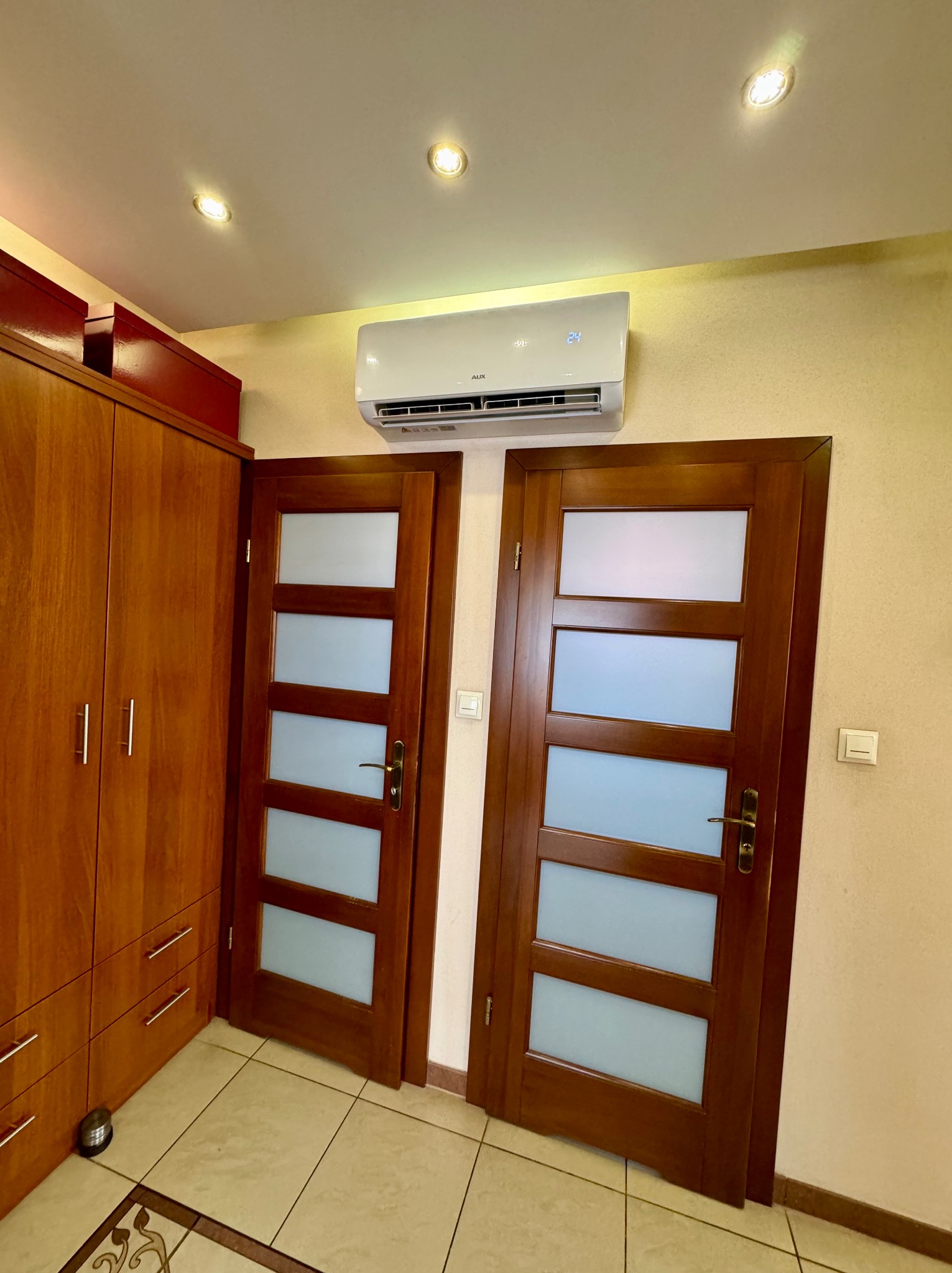 Biały klimatyzator marki AUX zamontowany nad dwuskrzydłowymi, drewnianymi drzwiami z mlecznym szkłem w mieszkaniu.