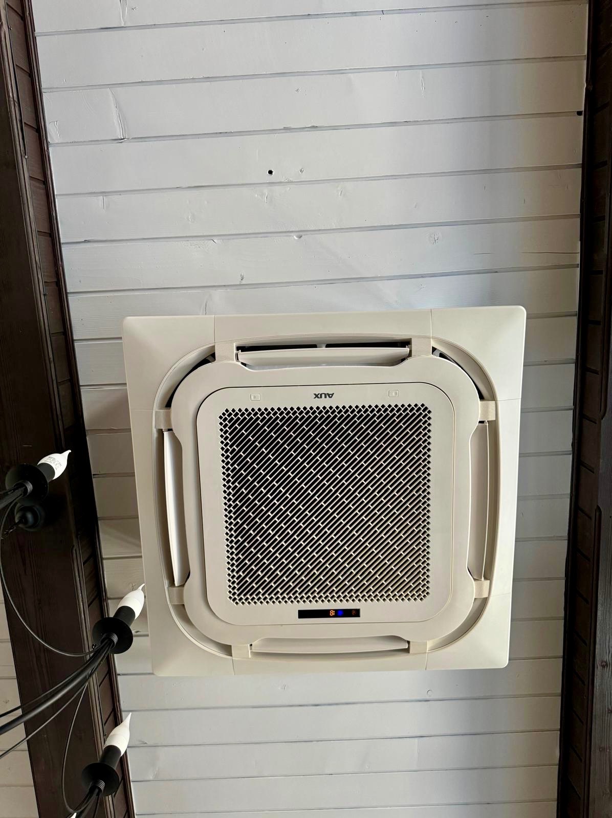 Kasetonowa klimatyzacja AUX zamontowana na białym, drewnianym suficie, widoczna z dołu, obok czarnego żyrandola.