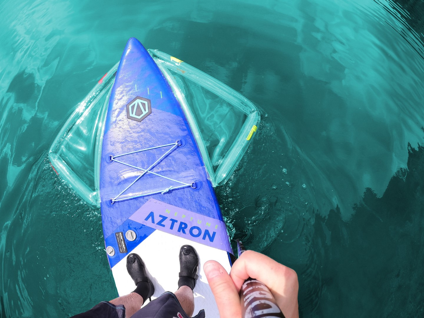 Osoba na niebiesko-białym paddleboardzie Aztron z przezroczystą dmuchaną platformą, wiosłuje po turkusowej wodzie, widok z góry.