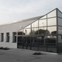 P.P.H.U. ERA-SZKŁO - Nowoczesny budynek z dużą, przeszkloną fasadą o nietypowym kształcie, z widocznymi szprosami i ciemną ramą, otoczony kostką brukową i niską zielenią.