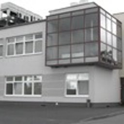 P.P.H.U. ERA-SZKŁO - Szary budynek biurowy z dużymi przeszkleniami okiennymi i balkonem z metalową konstrukcją, widok z dołu na tle pochmurnego nieba.