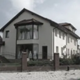 Dwukondygnacyjny dom z jasną elewacją, dużymi oknami z wykuszem i ciemną dachówką, otoczony niskim, ceglano-metalowym ogrodzeniem, widziany z perspektywy ulicy w pochmurny dzień.