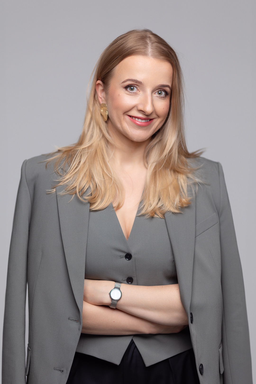 Portret uśmiechniętej blondynki w szarym garniturze z założonymi rękami, na neutralnym tle.