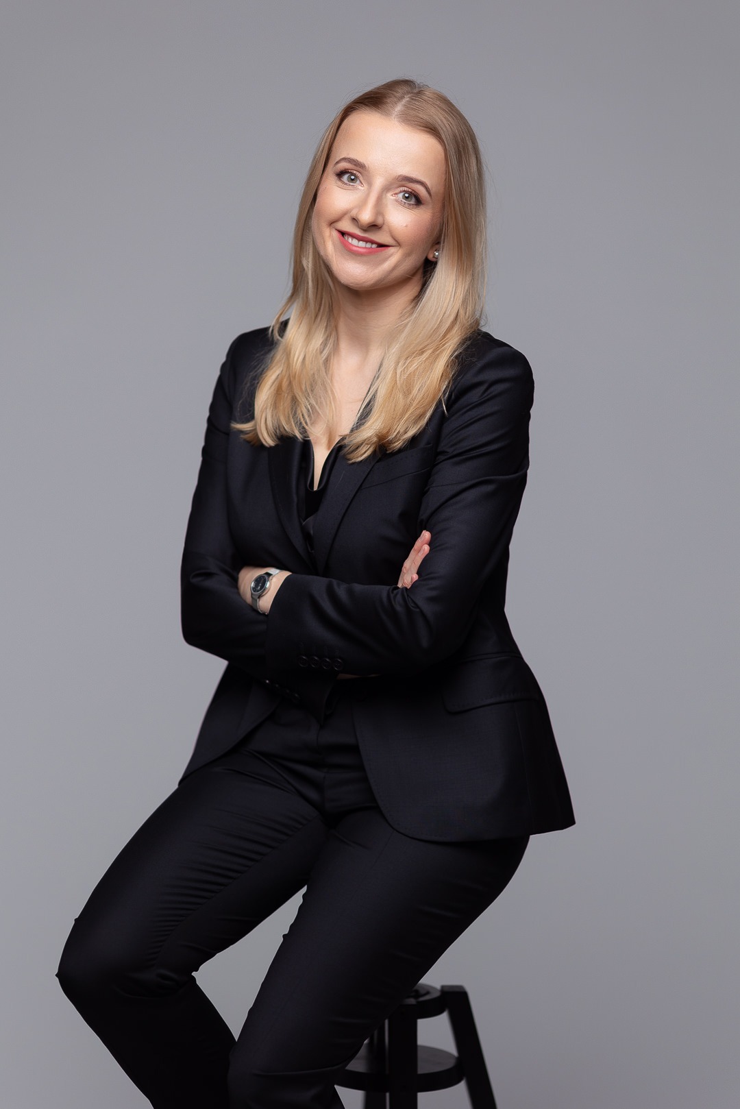 Portret uśmiechniętej blondynki w czarnym garniturze, siedzącej na stołku na szarym tle, z założonymi rękami i widocznym zegarkiem na nadgarstku.
