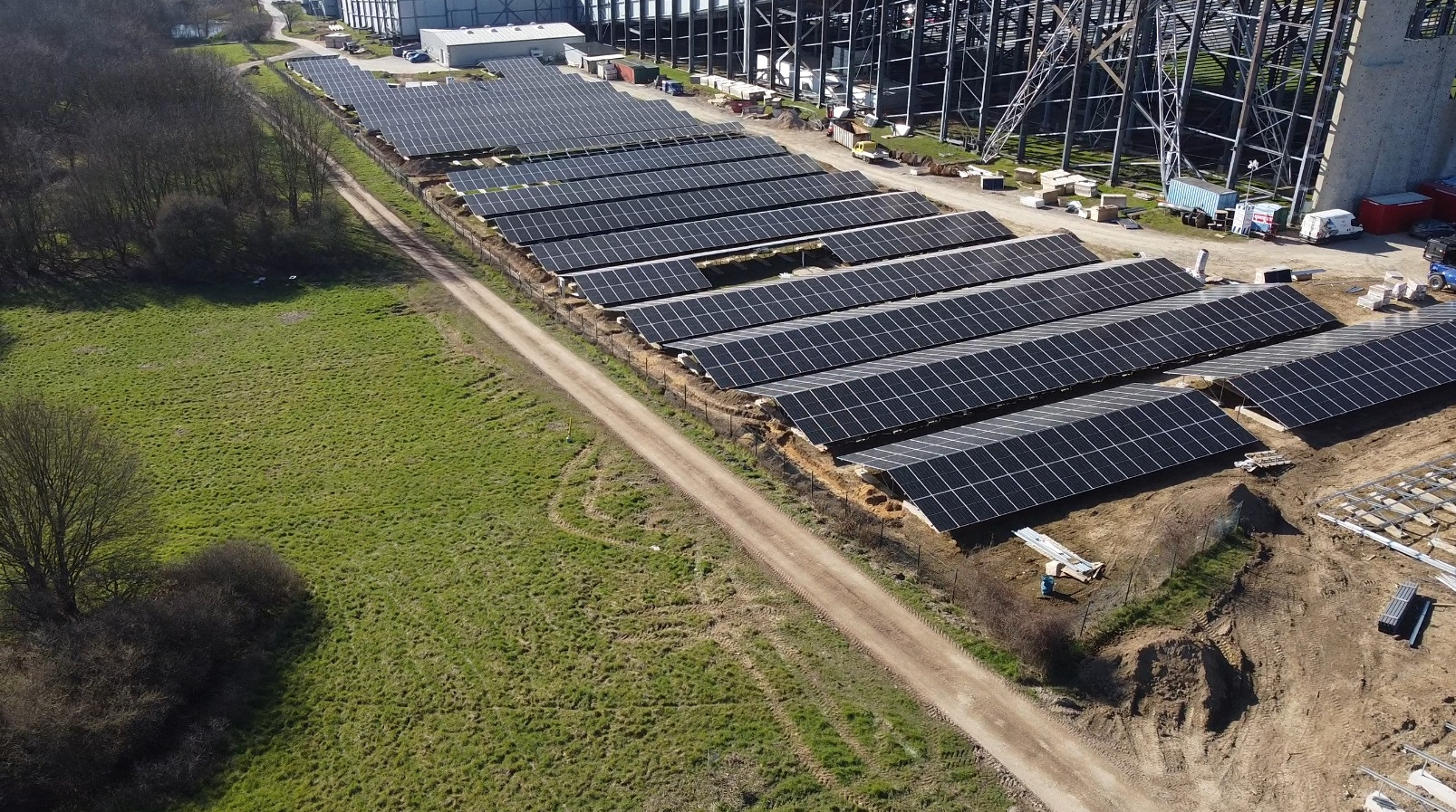 Rozległa instalacja paneli słonecznych na gruncie, widok z góry, obok konstrukcja stalowa hali, teren trawiasty i droga gruntowa.