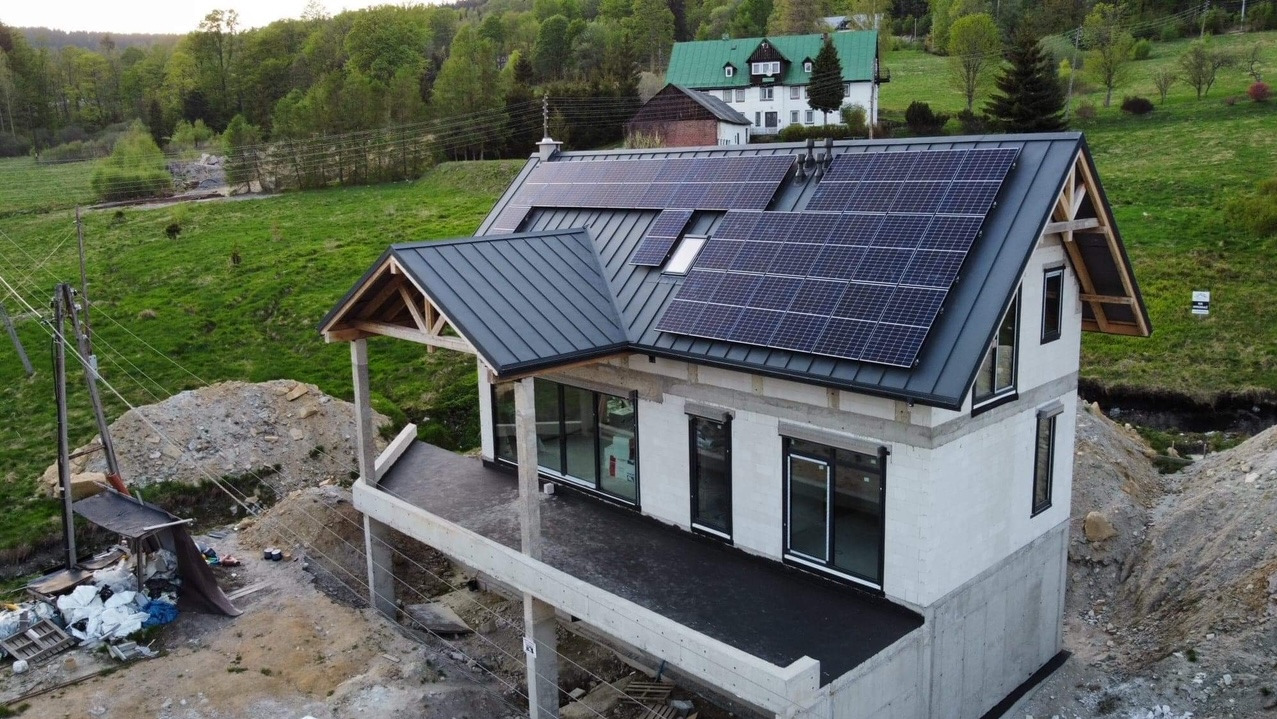 Niedokończony dom jednorodzinny z zainstalowanymi panelami fotowoltaicznymi na ciemnym dachu, widok z góry na tle zielonych wzgórz i sąsiedniego budynku.