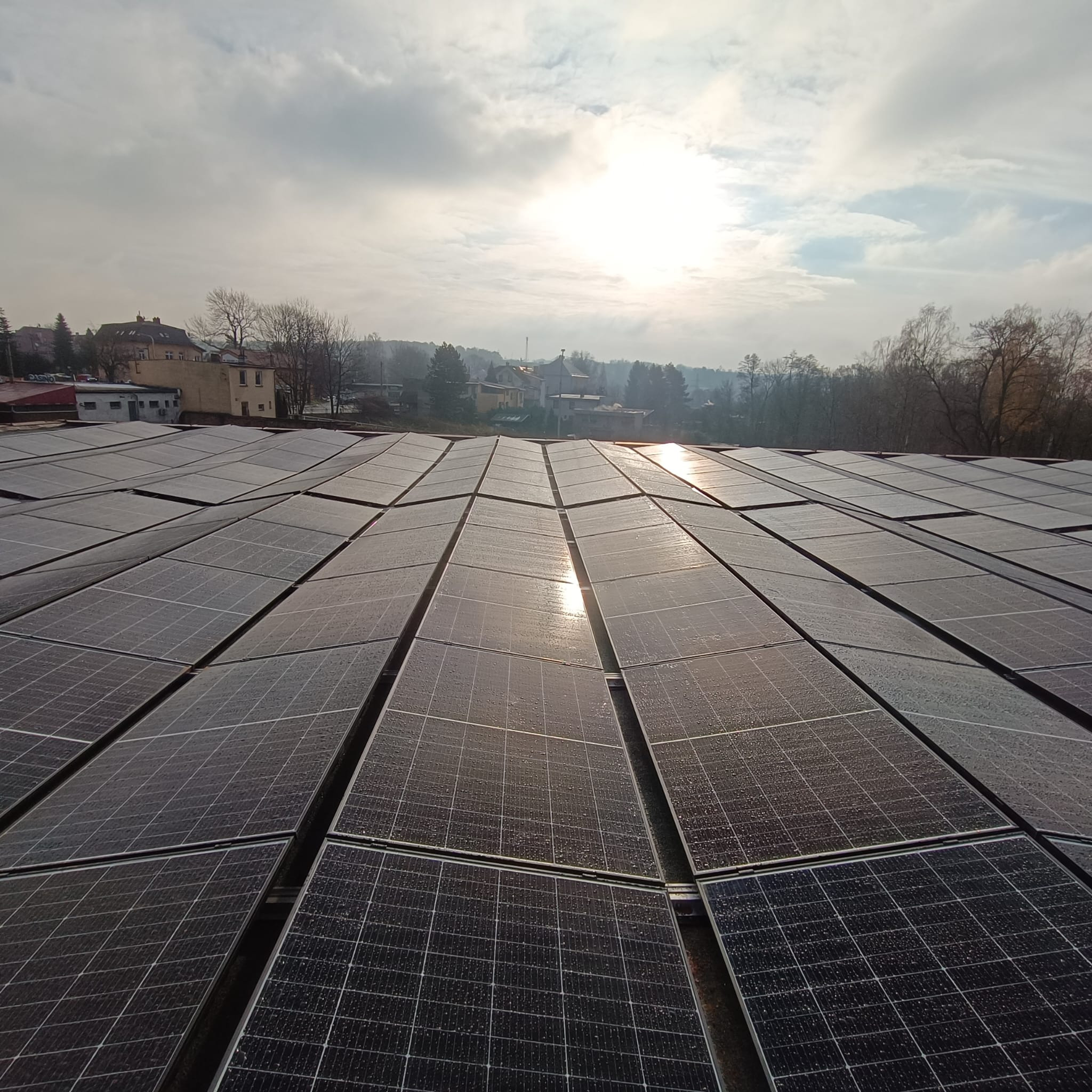 Rozległa instalacja fotowoltaiczna na dachu budynku w pochmurny dzień, z widocznymi kroplami wody na panelach i zabudowaniami w tle.