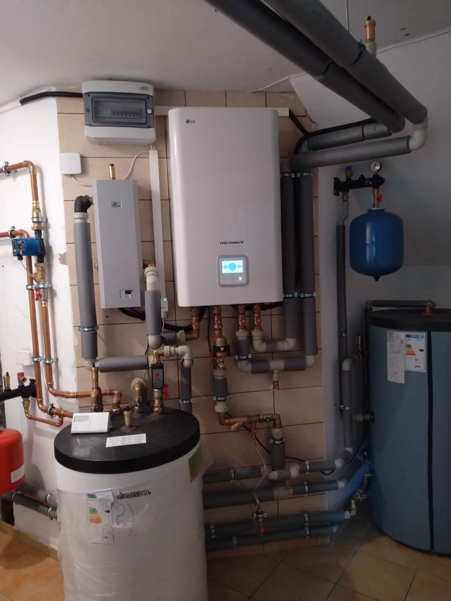 Zestaw urządzeń grzewczych: pompa ciepła LG Therma V, zasobnik ciepłej wody Kospel, naczynie przeponowe i rozdzielnica elektryczna na kafelkowanej ścianie.