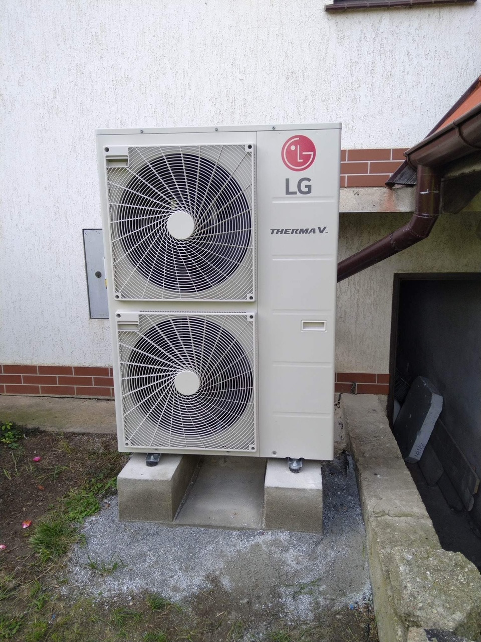 Zewnętrzna jednostka pompy ciepła LG Therma V zamontowana na betonowych blokach przy ścianie budynku z widoczną rynną.