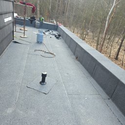 ROOF-TECH POLSKA PATRYK RATAJCZAK - Wykończony dach pokryty papą termozgrzewalną z kominkiem wentylacyjnym. Widoczne narzędzia i butle gazowe. Tło stanowią drzewa bez liści.