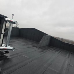 ROOF-TECH POLSKA PATRYK RATAJCZAK - Czarny, płaski dach pokryty papą termozgrzewalną z widoczną instalacją odgromową i elementami wentylacyjnymi. W tle pochmurne niebo i fragmenty zabudowy miejskiej.
