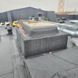 ROOF-TECH POLSKA PATRYK RATAJCZAK - Szare pokrycie dachu płaskiego z wyłazem dachowym i elementami wentylacji. W tle widoczny żuraw budowlany. Praca dekarska w trakcie realizacji.