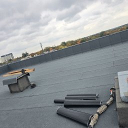 ROOF-TECH POLSKA PATRYK RATAJCZAK - Szary dach pokryty papą termozgrzewalną z kominem wentylacyjnym i przygotowanymi rurami do instalacji odgromowej na tle pochmurnego nieba i zabudowy miejskiej.