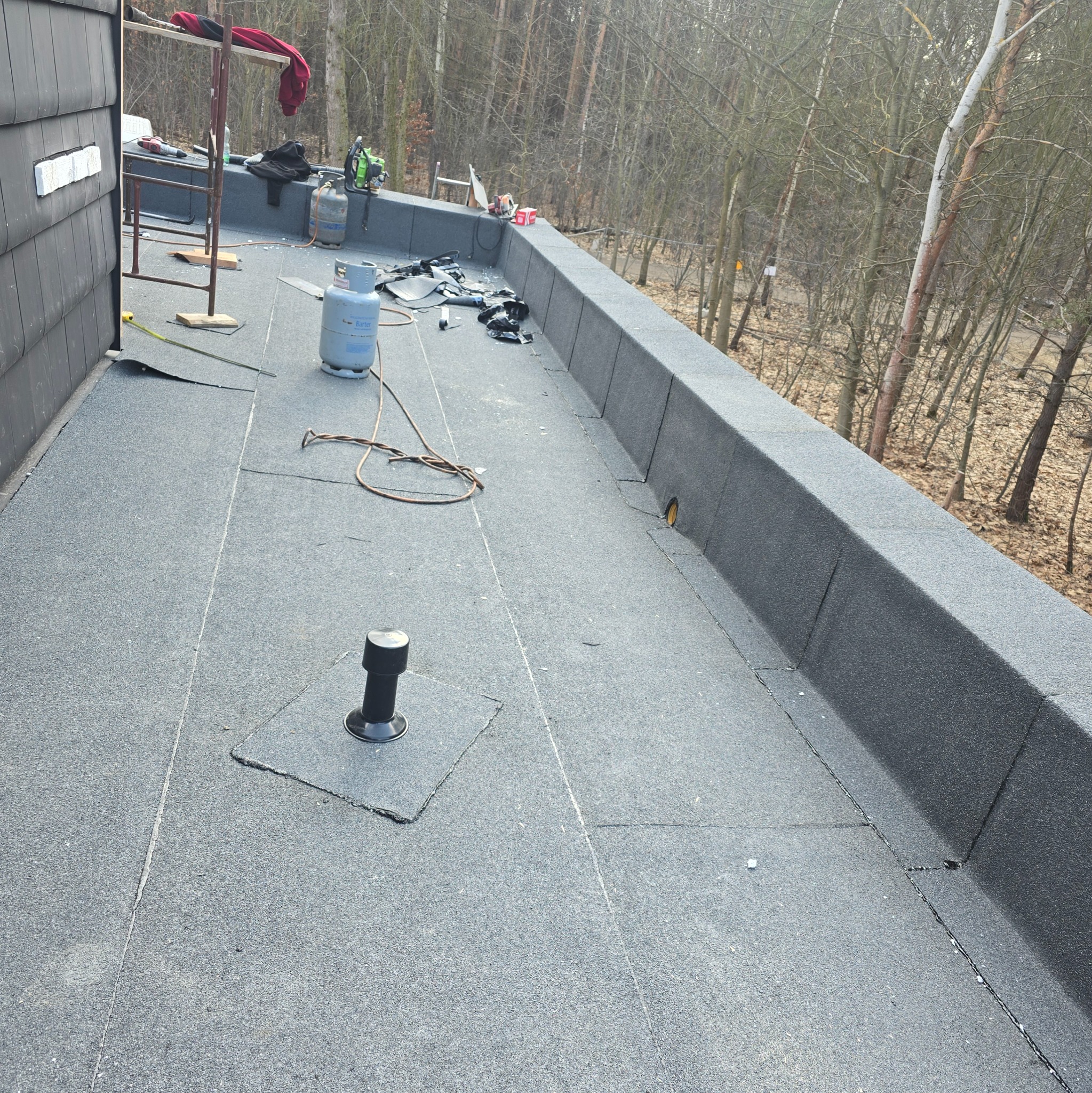 Wykończony dach pokryty papą termozgrzewalną z kominkiem wentylacyjnym. Widoczne narzędzia i butle gazowe. Tło stanowią drzewa bez liści.